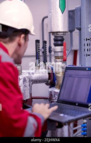 Thema der Energiewirtschaft: Ein Techniker in roter Kleidung und ein weißer Helm, der die Heizungsparameter auf einem tragbaren Computer überprüft. Stockfoto