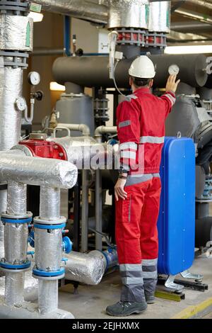 Thema der Energiewirtschaft: Ein Techniker in roten Overalls und einem weißen Helm, der die Heizungsparameter überprüft. Heizwasserverteilungstechnik. Stockfoto