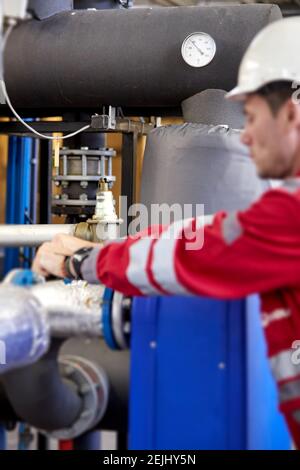 Thema der Energiewirtschaft: Ein Techniker in rotem Overall und ein weißer Helm, der die Heizungsparameter überprüft. Heizwasserverteilungstechnik. Stockfoto