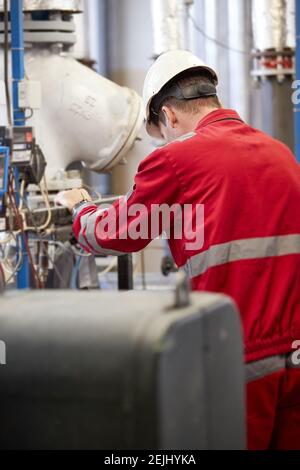 Thema der Energiewirtschaft: Ein Techniker in roten Overalls und einem weißen Helm, der die Heizungsparameter überprüft. Heizwasserverteilungstechnik. Stockfoto