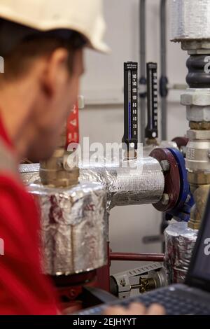 Thema der Energiewirtschaft: Ein Techniker in roten Overalls und einem weißen Helm, der die Heizungsparameter überprüft. Heizwasserverteilungstechnik. Stockfoto