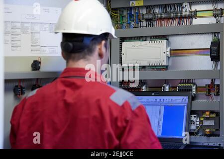 Thema der Energiewirtschaft: Ein Techniker in roten Overalls und einem weißen Helm, der die Heizungsparameter auf einem tragbaren Computer überprüft. Heizgeräte Stockfoto
