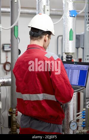 Thema der Energiewirtschaft: Ein Techniker in roten Overalls und einem weißen Helm, der die Heizungsparameter auf einem tragbaren Computer überprüft. Heizgeräte Stockfoto