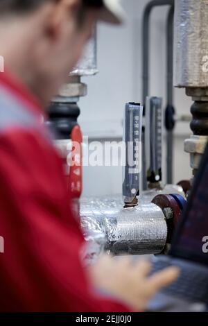 Thema der Energiewirtschaft: Ein Techniker in roten Overalls und einem weißen Helm, der die Heizungsparameter überprüft. Heizwasserverteilungstechnik. Stockfoto