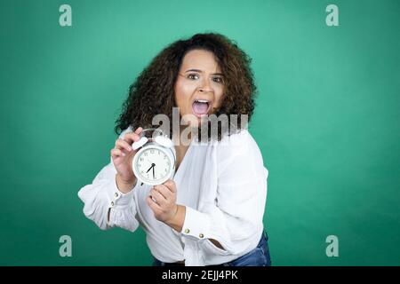 Junge afroamerikanische Mädchen trägt weißes Hemd über grünem Hintergrund schreiend und verängstigt, zeigt die Uhr Stockfoto