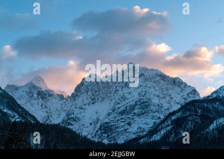 Wunderschöne Nahaufnahme eines rosa glühenden Berggipfeln in den Alpen bei Sonnenuntergang, während der Wind Schnee vom Berg weht. Kraft natürlicher Elemente Stockfoto