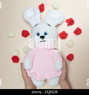 Handgemachtes Strickspielzeug. Gestricktes weißes Kaninchen in rosa Kleid in Händen auf hellem Hintergrund. Stockfoto