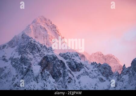 Wunderschöne Nahaufnahme eines rosa glühenden Berggipfeln in den Alpen bei Sonnenuntergang, während der Wind Schnee vom Berg weht. Kraft natürlicher Elemente Stockfoto