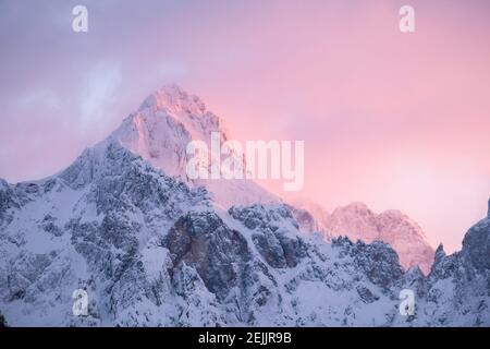 Wunderschöne Nahaufnahme eines rosa glühenden Berggipfeln in den Alpen bei Sonnenuntergang, während der Wind Schnee vom Berg weht. Kraft natürlicher Elemente Stockfoto