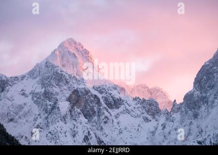 Wunderschöne Nahaufnahme eines rosa glühenden Berggipfeln in den Alpen bei Sonnenuntergang, während der Wind Schnee vom Berg weht. Kraft natürlicher Elemente Stockfoto