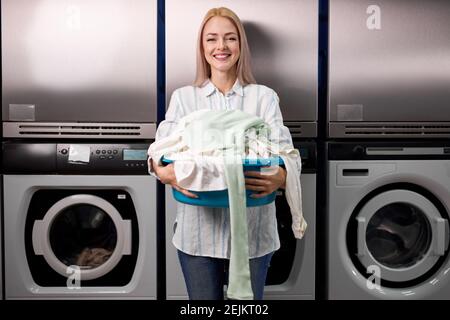 Junge Frau, die Wäsche im Waschsalon macht, schaut lächelnd auf die Kamera, hält Kleidung in den Händen und steht in der Nähe von Waschmaschinen. Waschen, putzen, laun Stockfoto