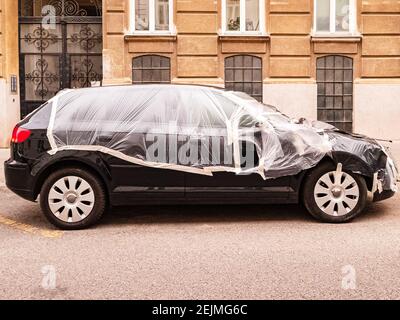 Schwarz Hatchback Auto ist mit transparentem Kunststoff-Wrap bedeckt und mit weißem Klebeband versiegelt. Steht auf der Straße vor dem Haus geparkt. Kaputt oder renoviert Stockfoto