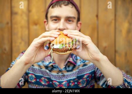 Kerl in einem stilvollen Shirt Kerl in einem stilvollen Shirt Mit einer Basiskappe genießen Sie einen schönen vegetarischen Burger auf Eine hölzerne Terrasse Stockfoto