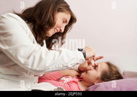 Ärztin lächelt und gibt einem kleinen Mädchen in ihrem Bett Augentropfen. Hausarzt Konzept Stockfoto