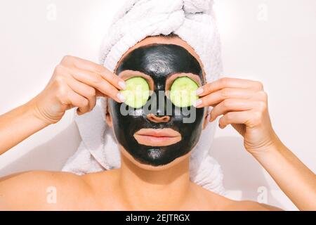 Frau mit reinigenden Holzkohle Gesichtsmaske hält Gurkenscheiben auf Ihre Augen Stockfoto