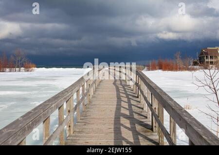 Eine alte Holzbrücke überquert einen Abschnitt der gefrorenen Georgian Bay Waterfront in Collingwood, Ontario. Der Himmel oben ist bedrohlich und düster, mit dem Winter Stockfoto