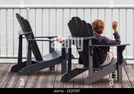 Erwachsene einsame Frau sitzt auf einem Holzstuhl auf dem Pier Blick auf den leeren Stuhl neben ihr. Traurigkeit und Trauer. Straßenansicht, selektiver Fokus. Stockfoto
