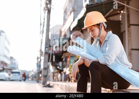 Architektur Frau müde und traurig und er sitzt auf der Boden und halten Blaupausen Design Plan und tragen Schutzhelm Auf der Baustelle in heißem w Stockfoto