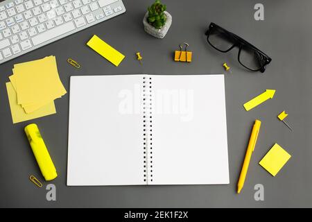 Geöffnetes Notebook und andere Bürogeräte wie Computertastatur, Bleistift, Aufkleber und Gläser auf dem hölzernen Büroschreibtisch. Gelbe und graue Farbe Stockfoto