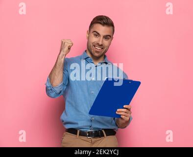 Aufgeregt junger Mann in Jeanshemd hält Clipboard, lacht und feiert Sieg mit Armen in der Luft, während posiert und auf rosa Hintergrund Stockfoto