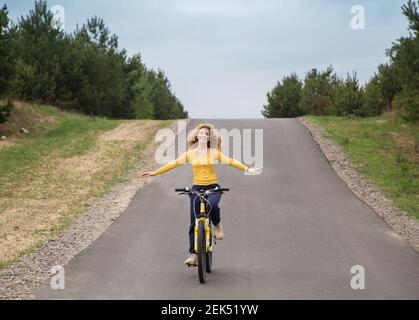 Nette fröhliche Teenager-Mädchen mit ausgestreckten Armen reitet ein Fahrrad auf der Straße vom Berg. Gehen Sie in der frischen Luft, Harmonie, Energie der Natur, rel Stockfoto
