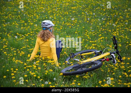 Unbekanntes Teenager-Mädchen in einem Fahrradhelm sitzt mit ihrem Rücken auf grünem Gras unter gelben Löwenzahn, Fahrrad liegt in der Nähe. Radfahren in der Natur, har Stockfoto