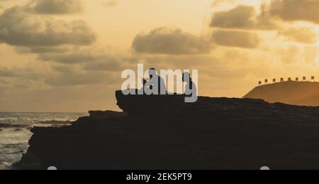 Sonnenuntergang über Accra Strand mit Felsen und zwei Personen sitzen Und genießen Sie das Meer mit hügeligen Boden, wo Sie sehen Die Schießstand Zeichen in der Hintergro Stockfoto