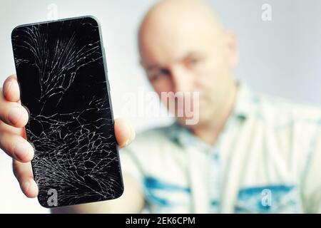 Telefon mit EINEM defekten Bildschirm Nahaufnahme. Ein weißer Mann hält ein schwarzes Smartphone mit einem geknackten Display. Stockfoto