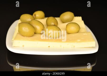 Mehrere würzige leckere Bio eingelegte grüne Oliven liegen auf Käse in Scheiben in einer weißen Keramikuntertasse. Der Hintergrund ist schwarz. Stockfoto