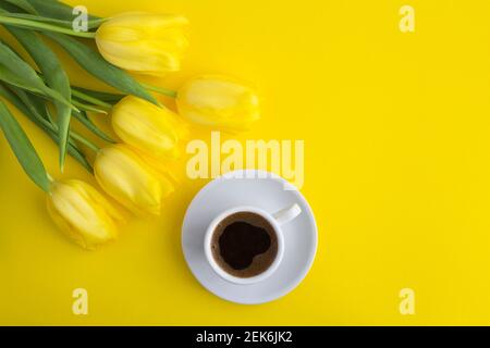 Schwarzer Kaffee in der weißen Tasse und gelbe Tulpen auf dem gelben Hintergrund.Draufsicht. Speicherplatz kopieren. Stockfoto