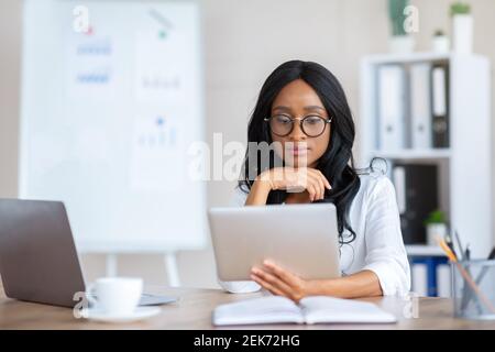 Fokussierte junge schwarze Geschäftsfrau mit Tablet-pc an ihrem Arbeitsplatz im modernen Büro, Kopierraum Stockfoto