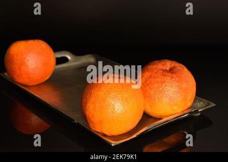 Drei organische reife saftige rote sizilianische Orangen auf einem Metalltablett. Der Hintergrund ist schwarz. Stockfoto