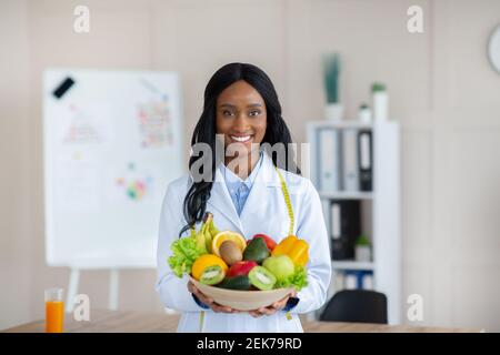 Porträt von glücklichen schwarzen Ernährungsberater im Labormantel hält Schüssel mit frischem Obst und Gemüse, lächelnd an der Kamera in der Klinik Stockfoto