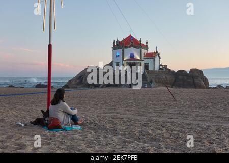 Kaukasische Frau mit Hund bei Sonnenuntergang in Miramar, Portugal, die wunderschöne Kapelle am Strand Capela do Senhor da Pedra Stockfoto