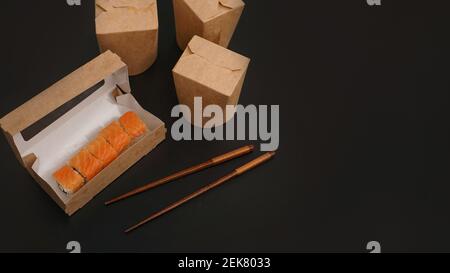 Lieferung asiatischer Speisen. Verpackung für Sushi und Woks. Lebensmittel in Papierbehältern auf schwarzem Hintergrund. Offenes Paket mit Lachsrollen Stockfoto