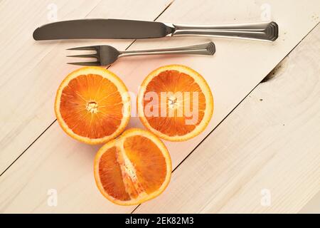 Drei Hälften eines reifen saftigen roten sizilianischen orange und Metall Besteck Messer und Dessertgabel, auf einem Hintergrund aus Naturholz weiß lackiert. Stockfoto