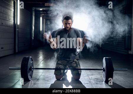 Bild von starken sportlichen Bodybuilder bereitet ihn selbst für schwere Heben. Kniend vor schwerem Gewicht im dunklen Raum. Stockfoto