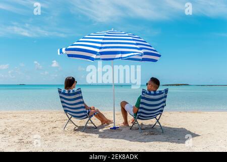 Ein glückliches Paar sitzt auf Stühlen unter dem Sonnenschirm an einem tropischen Strand. Sommerurlaub Konzept. Stockfoto