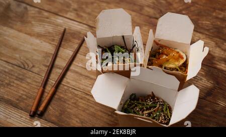 Nudeln mit Schweinefleisch und Gemüse in der Take-out-Box auf Holztisch. Lieferung asiatischer Speisen. Lebensmittel in Papierbehältern auf Holzhintergrund Stockfoto
