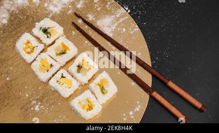 Sushi-Lieferung. Süße Brötchen aus Reis, Ananas, Kiwi und Mango. Rollt auf einem goldenen und schwarzen Hintergrund. Holzstöcke für Sushi. Stockfoto