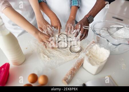 Ausschneiden von runden Formen aus gerolltem Teig, um hausgemachte Cookies vorzubereiten. Hände der multiethnischen Familie schneiden rohen Teig mit Metallschneider Stockfoto