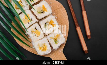 Dessert Sushi. Süße Kiwi, Ananas-Sushi-Brötchen. Sushi auf einem Holztablett auf schwarzem Hintergrund mit einem tropischen Blatt Stockfoto