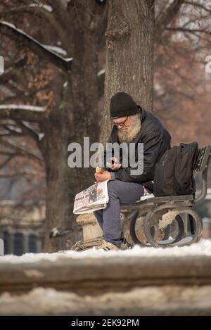 Prag, Tschechische Republik. 02-23-2021. Der alte Obdachlose isst und liest die Zeitung auf einer Bank im Stadtzentrum von Prag auf einem kalten Sieg Stockfoto