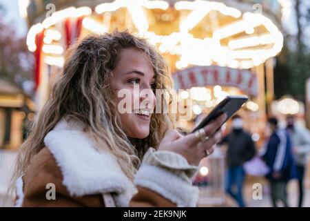 Junge Frau, die im Stehen Sprachnachrichten über das Smartphone sendet Gegen beleuchtetes Karussell Stockfoto