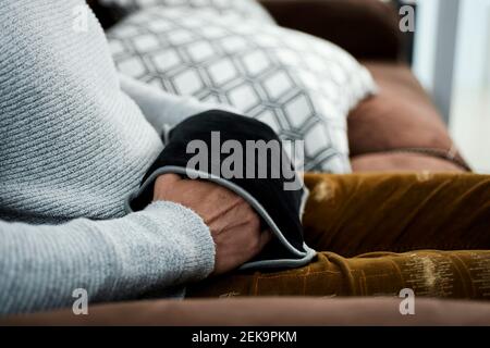 Ein junger Kaukasusmann benutzt einen schwarzen elektrischen Heißwasserbeutel, um seine Hände zu wärmen, während er auf einer bequemen braunen Couch sitzt Stockfoto