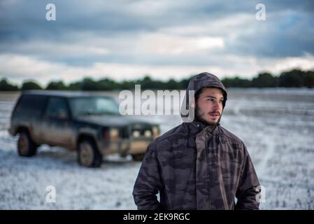 Junger Mann in Kapuzenjacke, der bei der Landwirtschaft gegen das Auto steht Ein Stockfoto