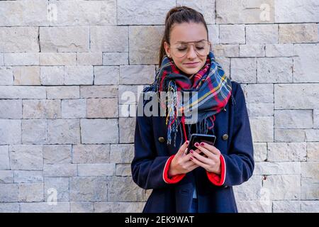 Teenager-Mädchen tragen warme Kleidung mit Handy im Stehen Gegen Backsteinmauer zu Hause Stockfoto