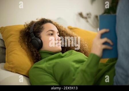 Junge Frau mit Buch liegt auf dem Bett, während Musik zu hören Über Kopfhörer zu Hause Stockfoto