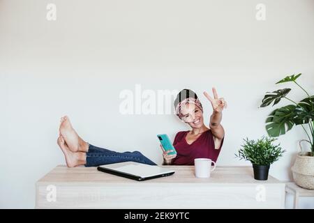 Fröhliche Unternehmerin, die Friedenszeichen macht, während sie sich mit den Füßen entspannt Im Home Office Stockfoto