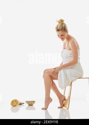 Frau im Handtuch Anwendung Feuchtigkeitscreme auf Bein, während sitzend auf Stuhl vor weißem Hintergrund Stockfoto